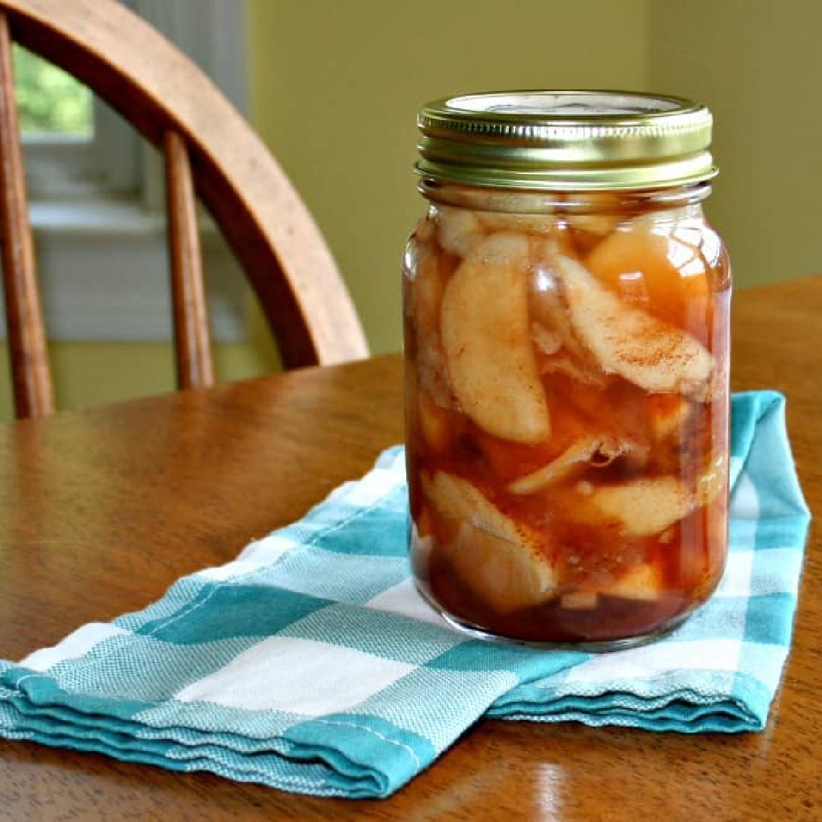 a jar of canned apples on a blue checked napkin sitting on a wooden kitchen table